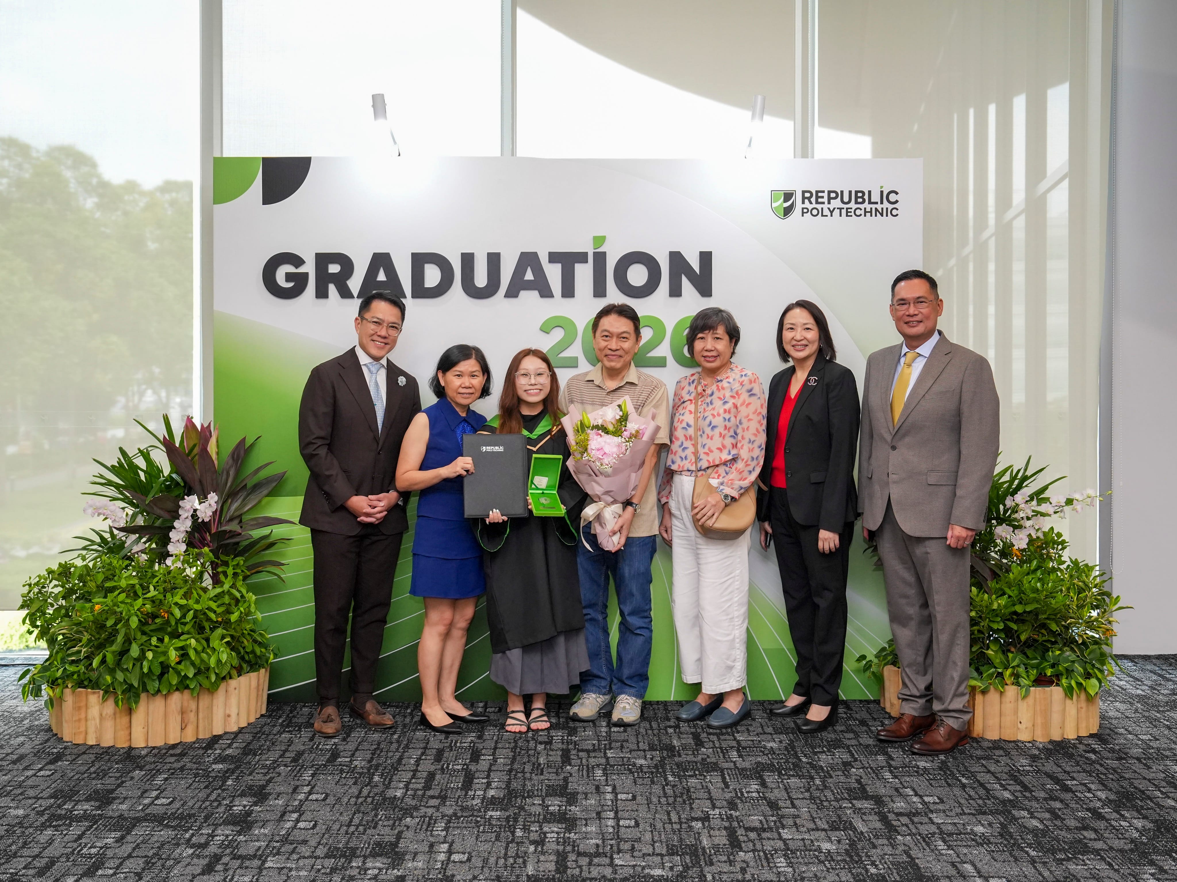 Group photo at a Republic Polytechnic graduation event. A graduate in academic gown stands at the centre holding a diploma folder and a medal case, with a bouquet of flowers, surrounded by six adults. The group poses in front of a large backdrop displaying “GRADUATION 2026” and the Republic Polytechnic logo, with decorative plants on both sides.